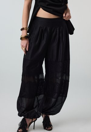 Frau trägt lockere schwarze transparente Hosen mit Spitzenpaneelen und hochhackigen Fransen-Sandalen, dazu Schmuck wie Armbänder und Ringe.