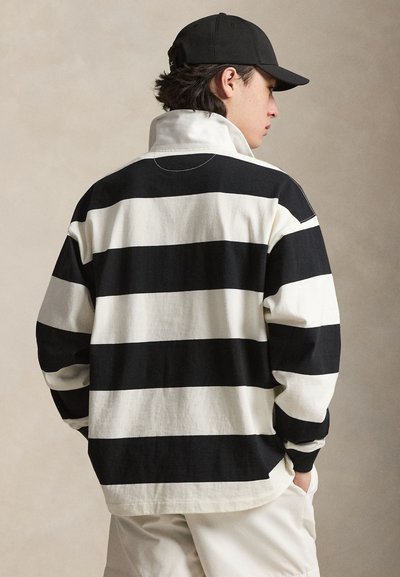 Persoon die een zwart-wit horizontaal gestreept rugbyshirt, een zwarte pet en witte broek draagt, met de rug naar de camera en handen in de zakken.