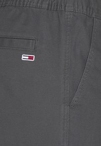 Gros plan d'un pantalon gris foncé avec ceinture élastique, poche arrière, couture latérale, et un petit patch logo rectangulaire rouge, blanc et noir.