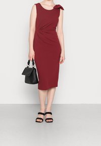 Robes sans manches bordeaux avec un détail torsadé sur le côté et un accent en nœud, associées à un sac noir et des sandales noires à lanières. Texture de tissu lisse.