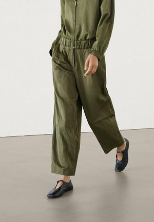 Person trägt olivgrüne, weit geschnittene Hosen und eine passende Jacke, geht auf einem ebenen Boden und trägt schwarze Mary Jane Schuhe.