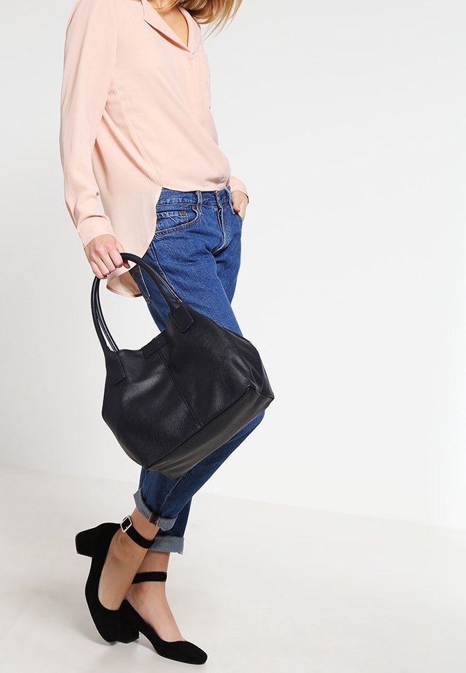 Femme portant un jean bleu retroussé, une blouse rose pâle, des escarpins noirs à bride cheville, tenant un grand sac à main en cuir noir avec deux anses.