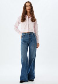 Camisa cropped de botones en rosa claro con detalles de lazo en las mangas, combinada con jeans acampanados de tiro alto en azul, sobre un fondo neutro.
