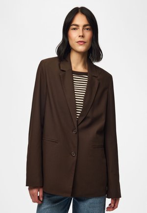 Femme aux cheveux foncés mi-longs portant un blazer marron foncé sur une chemise rayée noire et blanche et un jean bleu.