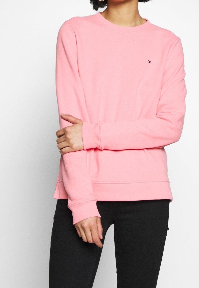 Mujer vestida con un sudadera rosa de manga larga con un pequeño logo en el pecho y pantalones negros, con los brazos cruzados al frente.