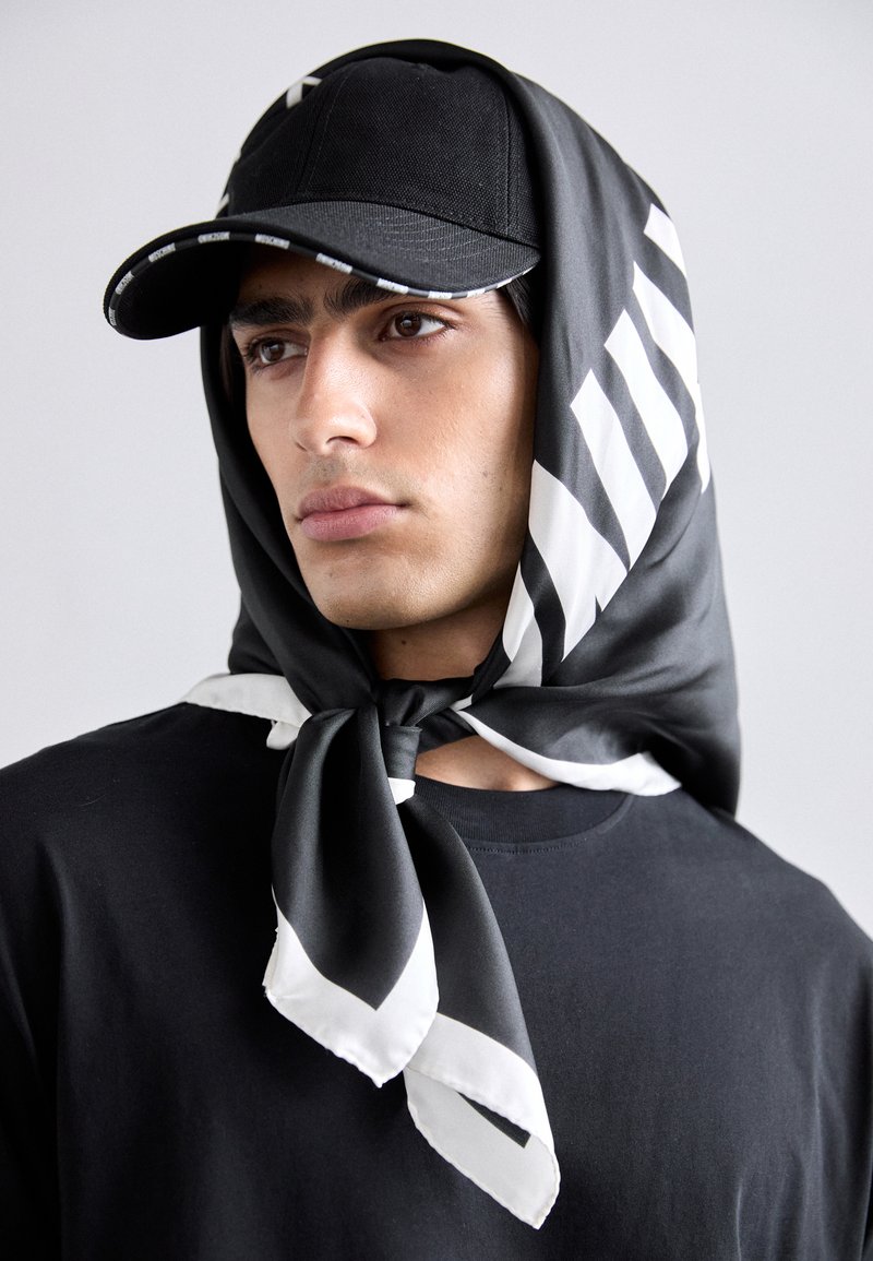 Sort tørklæde bundet om halsen med hvide diagonale striber, parret med en sort hat med mønstret skygge, båret over en sort skjorte.
