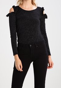 Černý dlouhý rukáv top s výřezem na ramenech, texturovaný materiál a volánové akcenty na ramenou. Spárován s černými skinny džínami.