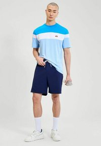 Jonge man staat, draagt een blauw en wit sport T-shirt, marineblauwe shorts, witte sokken en sneakers, en houdt een heldere waterfles in zijn rechterhand.