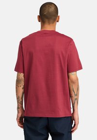 Camiseta corta de manga en burdeos con cuello redondo. Presenta un corte relajado y una tela suave. Muestra brazos tatuados sobre un fondo liso.