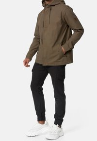 Chaqueta verde oliva con capucha, cremallera frontal y bolsillos laterales. Combinada con pantalones cargo negros y zapatillas blancas. Diseño simple y funcional.