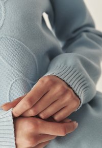 Hele sinine kudumipullover, millel on soonikust mansetid ja tekstureeritud muster, millel on peened kõverad, esitades pehme ja siledat kangast.