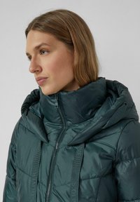 Napięta, ciemnozielona kurtka puchowa z wysokim kołnierzem, z zamkiem błyskawicznym i markowymi paskami na przodzie. Gładka, błyszcząca tekstura.