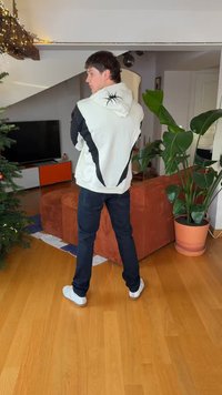 Balta kapuce džemperis ar melniem akcentiem un zirneklim līdzīgu grafiku uz muguras, kopā ar tumšām džinsu biksēm un baltām kurpēm, stāvot viesistabā.