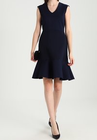 Mörkblå klänning med V-ringning, ärmlös design och utsvängd kjol. Tyget verkar vara texturerat, och en svart kuvertväska hålls i vänster hand.