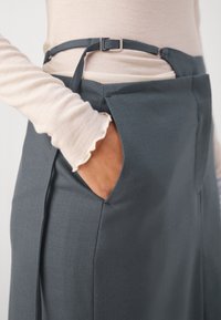 Dłoń w kieszeni ciemnoszarej spódnicy z wysokim stanem i regulowanym paskiem, zestawiona z jasnobeżowym, prążkowanym topem z długim rękawem i falbankowymi mankietami.