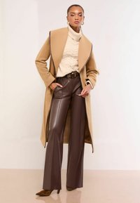 Kamel lang frakke med mørke kanter, cremefarvet kabelstrik turtleneck sweater, brune faux læder brede bukser og spidse brune hæle.