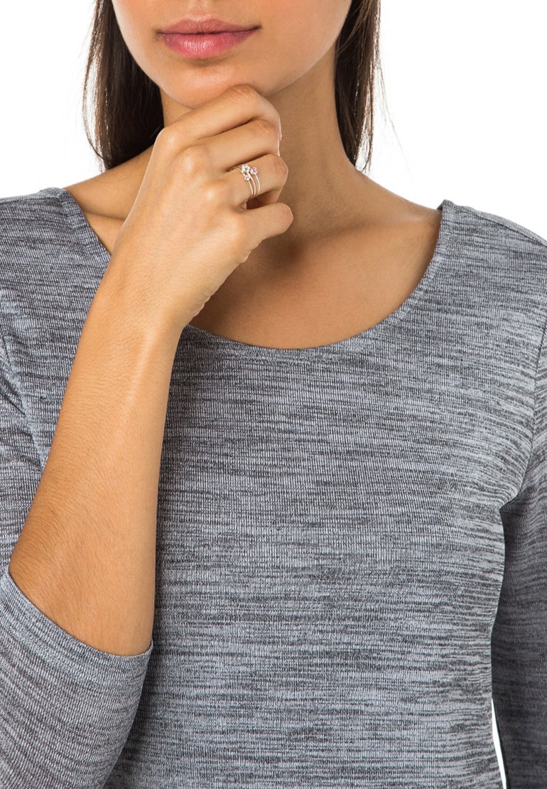 Grå, struktureret langærmet top. En tre-bånd ring med en lille pink ædelsten hviler på modellens hånd, som blidt berører hendes hage.