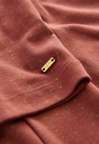 Bordeaux Baumwollstoff mit feinem Muster aus gelben Punkten, versehen mit einem kleinen goldenen Etikett mit der Aufschrift "NEXT", das in die Seitennaht genäht ist.