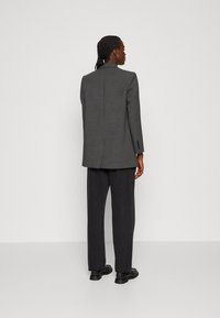 Grijze blazer met een gestructureerd ontwerp, enkele split aan de achterkant en knoopmanchetten, gecombineerd met zwarte wijde broek en zwarte schoenen.