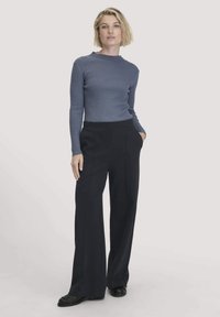 Gestreifter, langärmliger Pullover in Blau, kombiniert mit hoch taillierten, weit geschnittenen schwarzen Hosen. Das Model steht mit einer Hand in der Tasche und trägt schwarze Schuhe.