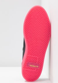 Black sneaker sole with a textured pattern; bright pink rubber outsole featuring a yellow-gold logo patch near the heel.