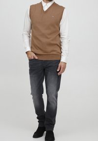 Brun ärmlös tröja med V-ringning, bärs över en vit långärmad skjorta. Kombinerad med mörkgrå jeans och svarta skor.