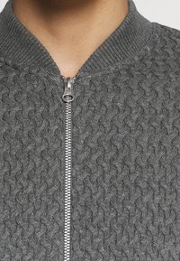 Grå stickad bomberjacka med ett texturerat mönster, har en framsidans dragkedja och ribbad krage, vilket betonar värme och komfort.