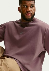 Άνδρας με κοντή γενειάδα, φοράει φούτερ Nike σε μωβ απόχρωση και μπεζ παντελόνι, κοιτάζει δεξιά πάνω σε απλό ανοιχτό φόντο.