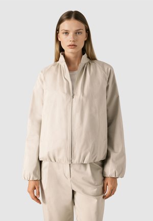 Kobieta z prostymi włosami ubrana w beżową kurtkę na zamek i pasujące spodnie, stoi na tle jednolitego jasnoszarego tła.