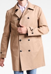 Beige trenchcoat i en bomullsblandning, med dubbelknäppt design, svarta knappar och sidofickor, som bärs över en vit skjorta.