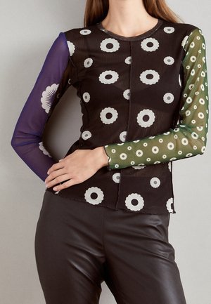 Femme portant un haut noir transparent avec des motifs de marguerites blanches et des manches longues dépareillées en violet et vert, associé à un pantalon en cuir foncé.