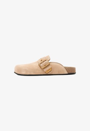 Beige ruskind slip-on træsko med stort justerbart guldbeslag og korksål, designet til afslappet brug.