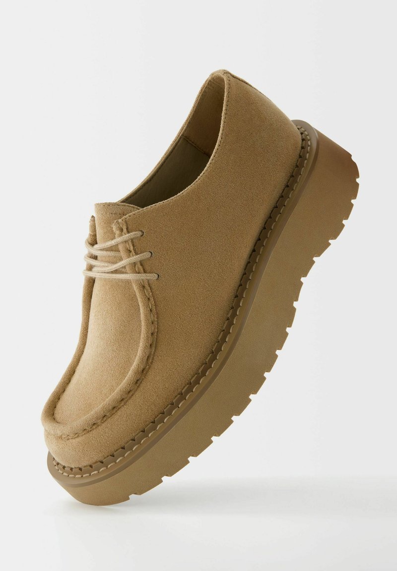 Zapatos de ante beige con punta redonda, completados por una suela de goma gruesa y texturizada, con un mínimo detalle de cordones en la parte delantera.