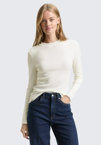 Jonge vrouw met lang blond haar draagt een wit shirt met lange mouwen en donkerblauwe jeans met hoge taille, met één hand in de zak.