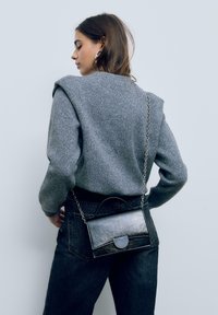 Borsa a tracolla in pelle nera con manico arrotondato, forma rettangolare, tracolla a catena e chiusura a pattina curva. Indossata su un maglione grigio.