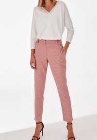 Haut blanc à col en V et manches longues avec une coupe décontractée associé à un pantalon ajusté à carreaux rouges et blancs. Modèle portant des sandales à talons aiguilles noires.