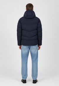 Granatowa pikowana kurtka puchowa z przeszyciami w kształcie litery V, z kapturem i ocieplanym krojem, noszona na jasnoniebieskich dżinsach i czarnych butach.