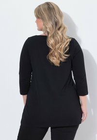 Czarna tunika z rękawami do łokcia, okrągłym dekoltem, miękką tkaniną i lekko zaokrąglonym dołem, w zestawie z czarnymi spodniami.