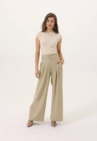 Bež rebrasti top i hlače maslinasto zelene boje visokog struka s širokim nogavicama. Glatka tkanina s naborima; model nosi bež štikle i minimalne dodatke.