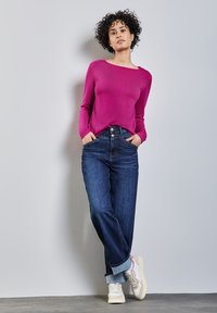 Fuchsia Strickpullover mit weitem Ausschnitt, kombiniert mit locker sitzenden dunkelblauen Jeans und weißen Sneakers mit beige Akzenten. Das Model steht an einer Wand.