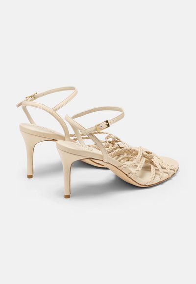 Sandales à talons hauts beiges avec des brides tressées sur les orteils, de fines brides de cheville et un détail de boucle en ton doré. Texture en cuir lisse, design à talon étroit.