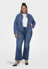 Denim jakna temno modre barve s sproščenim krojem, dvema žepoma na prsih in enobarvno belo srajco. V kombinaciji z razširjenimi modrimi kavbojskimi hlačami in petkami.