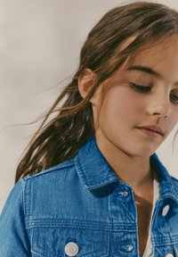 Chaqueta de mezclilla azul con cierre de botones, cuello clásico y detalles de textura; llevada por una chica con cabello suelto y ondulado.