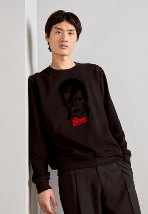 Sudadera negra con un gráfico de una cara estilizada con un rayo y el texto "Bowie" en rojo. Tejido suave con puños acanalados.