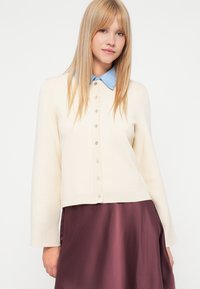 Krém színű kardigán világoskék gallérral, gombos elülső résszel és bordás mandzsettákkal, sötét szilva szoknya fölött.