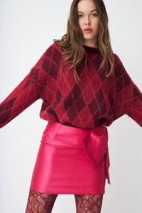 Kobieta w dużym czerwono-bordowym swetrze w romby, różowej satynowej mini spódnicy z kokardą oraz bordowych koronkowych rajstopach na jednolitym tle.