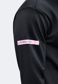 Gros plan sur la manche d'une chemise noire à manches longues avec un patch rectangulaire rose indiquant "ENDLESS" et comportant un symbole infini.