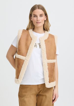 Femme portant une veste en peau lainée beige par-dessus un t-shirt blanc et un pantalon marron, debout devant un fond clair uni.