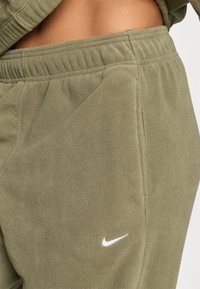 Olijfgroene fleece joggers met een elastische tailleband, voorzien van een klein wit Nike-logo op de voorkant van de broekzak voor branding. Zachte textuur.