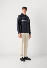 Hanorac de marină cu logo texturat "BOSS" și dungi gri, asortat cu pantaloni crem și adidași negri. O geantă atârnă de umărul stâng.